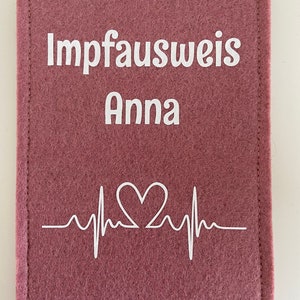 Könnte beinhalten: Eine rechteckige Hülle aus altrosa Filz mit weißem, aufgedrucktem Text Impfausweis Anna über einer weißen Grafik einer Herzschlaglinie, die in der Mitte ein Herz bildet. Die Hülle ist an den linken und rechten Seiten mit Nähten versehen.