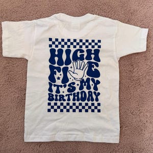 Könnte beinhalten: Ein weißes kurzärmeliges T-Shirt mit einem dunkelblauen Grafikdruck. Das Design umfasst einen Schachbrettmuster-Rand oben und unten, den Text "HIGH FIVE IT'S MY BIRTHDAY" in einer wellenförmigen Schrift und eine zentrale Abbildung einer offenen Hand.