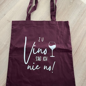 Könnte beinhalten: Eine bordeauxfarbene Tragetasche mit weißem Text, der "Zu Vino Sag Ich Nie No!" lautet, und einer weißen Umrandung eines Weinglases.