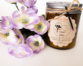 Puede incluir: Un tarro de vidrio marrón con una vela, una tapa plateada y una etiqueta de papel marrón. La etiqueta tiene un dibujo de un árbol Joshua y el texto "Love Spell Desert Wicks Batch No. 2 Pour No. 1 Handcrafted Natural Soy Candle Net WT 8 oz".