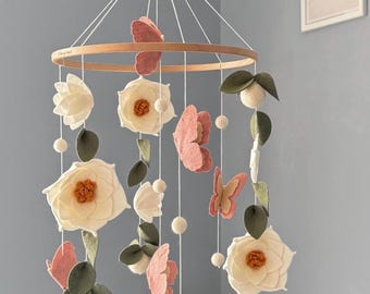 May include: A hanging mobile featuring a circular wooden hoop suspended by white strings. Attached are felt elements including white flowers with orange centers, pink butterflies, dark green leaves, and small white felt balls hanging at various lengths.