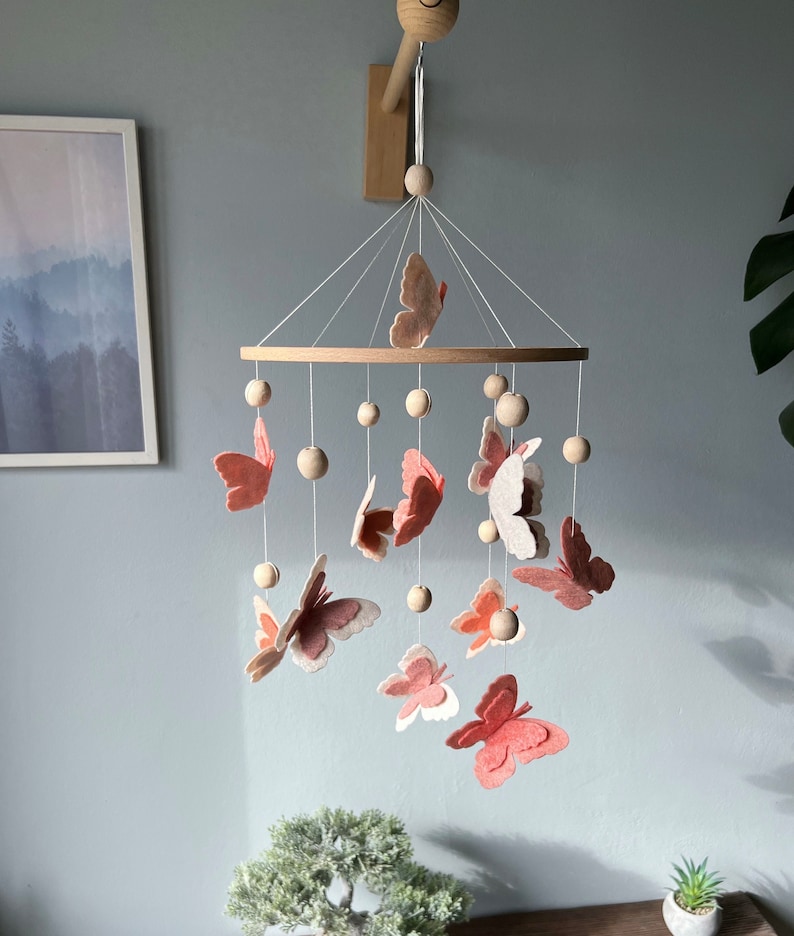 May include: A wooden mobile with pink and white felt butterflies and wooden beads hanging from a circular wooden frame. The mobile is suspended from a wooden hook attached to the ceiling.
