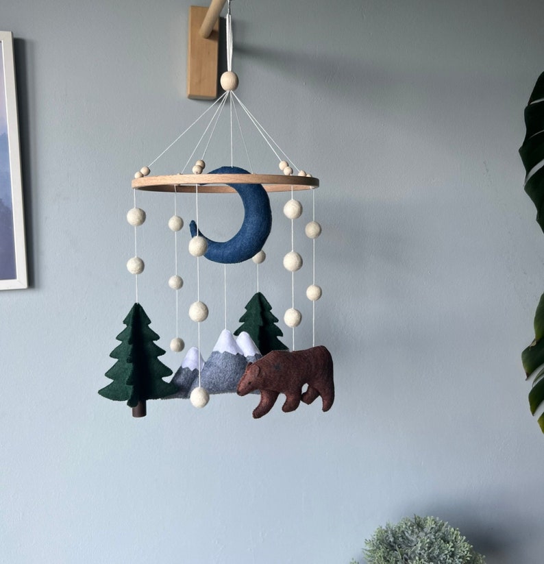 May include: A felt baby mobile with a blue crescent moon, white pom-poms, green trees, gray mountains, and a brown bear.