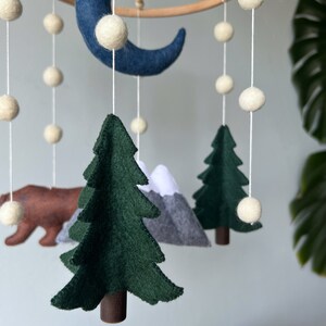 May include: A felt baby mobile with a blue crescent moon, white felt balls, green felt trees, a brown felt bear, and gray felt mountains.