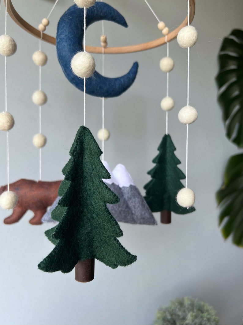 May include: A felt baby mobile with a blue crescent moon, white felt balls, green felt trees, a brown felt bear, and gray felt mountains.