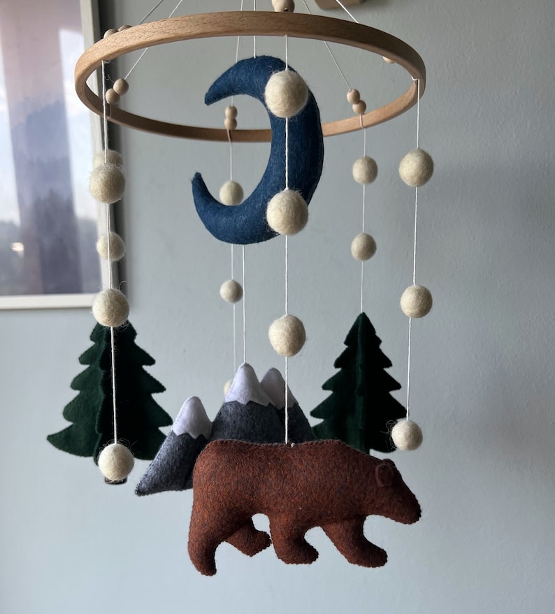 May include: A felt baby mobile with a brown bear, green trees, gray mountains, a white crescent moon, and white pom-poms hanging from a wooden ring.