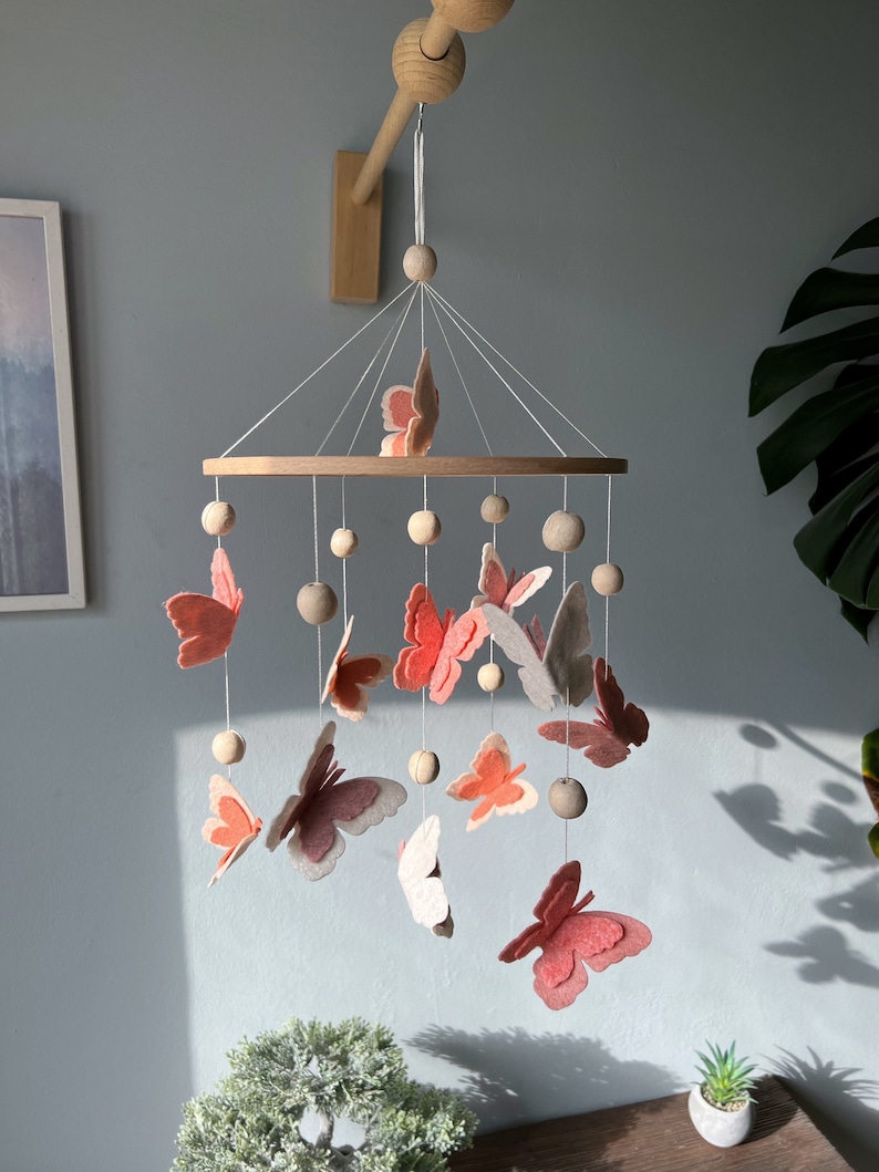 May include: A wooden baby mobile with a circular wooden frame and pink and white felt butterflies hanging from it. The mobile is suspended from a wooden rod attached to the ceiling.