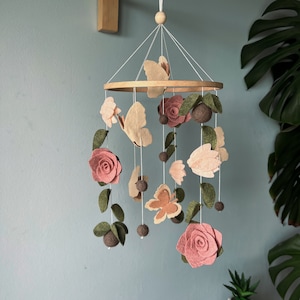 Puede incluir: Un móvil de bebé de madera con flores, mariposas y hojas de fieltro en tonos rosa, marrón y verde. El móvil está colgado de un brazo de madera sujeto a la pared.