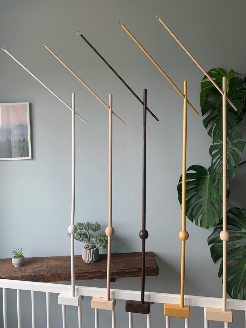 May include: Six wooden mobile arm stands in different colors, including white, beige, brown, black, yellow, and natural wood. Each stand has a round wooden ball at the top and a wooden base. The stands are mounted on a white railing.