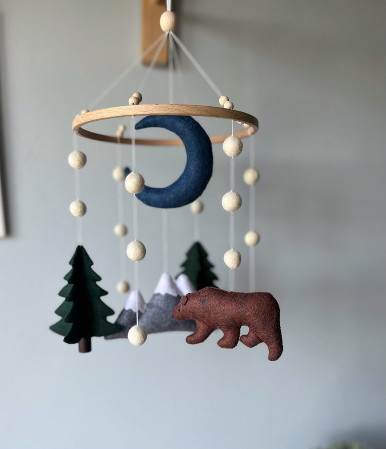 May include: A felt baby mobile with a blue crescent moon, a green pine tree, a brown bear, and gray mountains. The mobile is suspended from a wooden ring with white felt balls.
