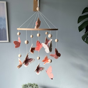 May include: A wooden baby mobile with a circular wooden frame and pink and white felt butterflies hanging from it. The mobile is suspended from a wooden hook.