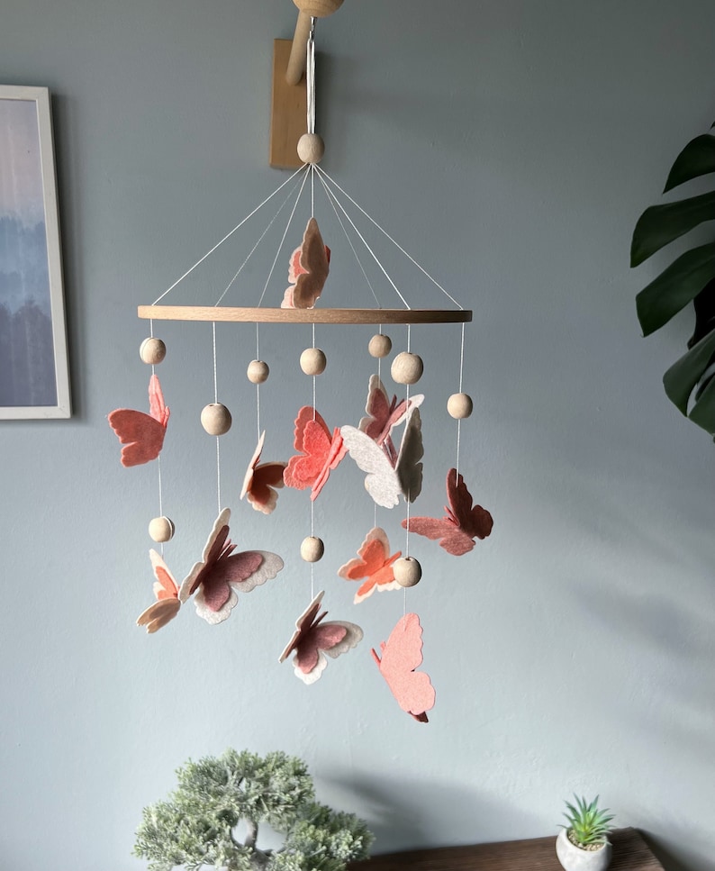 May include: A wooden baby mobile with a circular wooden frame and pink and white felt butterflies hanging from it. The mobile is suspended from a wooden hook.