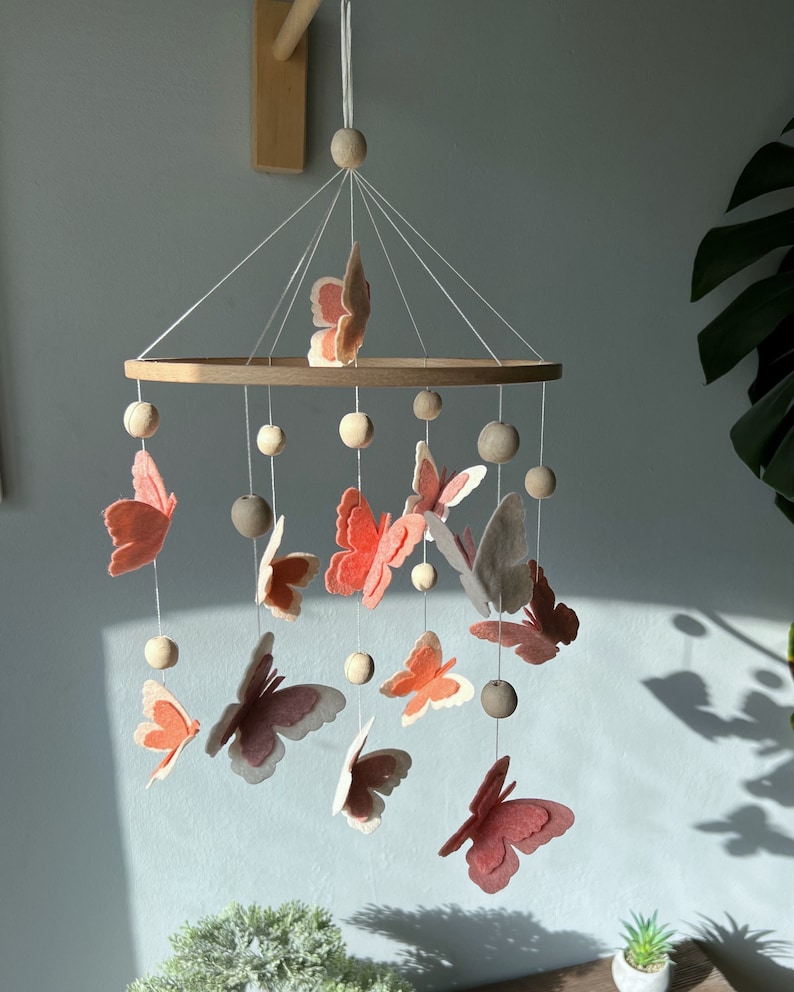 May include: A wooden baby mobile with a circular wooden frame and pink and white felt butterflies hanging from it. The mobile is decorated with wooden beads.