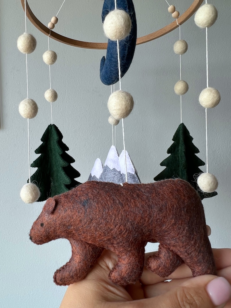 May include: A brown felt bear, two green felt trees, a gray felt mountain range, and white felt balls hanging from a wooden ring. The bear is the focal point of the image.