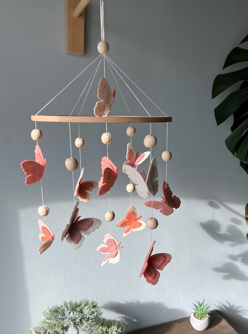May include: A wooden baby mobile with a circular frame and pink and white felt butterflies hanging from it. The mobile is decorated with wooden beads.