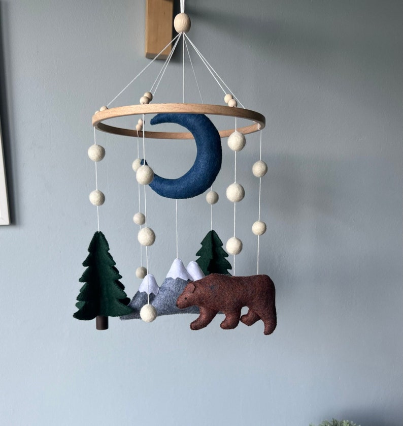 May include: A wooden mobile with felt woodland animals and a blue crescent moon. The mobile features a brown bear, two green trees, and gray mountains. White felt balls are strung throughout the mobile.