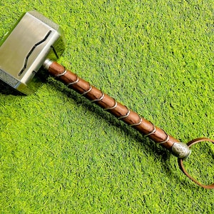 May include: A silver-colored hammer with a brown leather handle and a silver ring at the end. The hammer is on a green grassy surface.