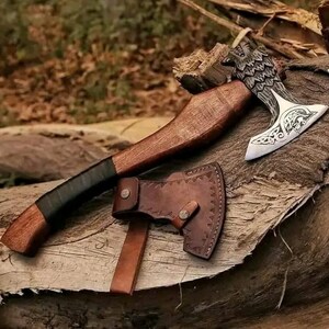 May include: A hand-crafted axe with a wooden handle and a leather sheath. The axe head features a detailed carving of a bird and intricate patterns.