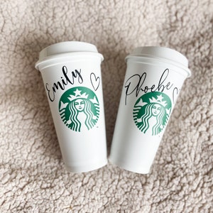 May include: Two white takeaway coffee cups with black lids. The cups feature a green Starbucks logo with a woman's face and the names "Emily" and "Phoebe" written on them.