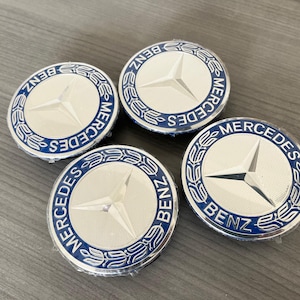 May include: Four silver and blue Mercedes-Benz wheel center caps. Each cap features the Mercedes-Benz logo, a three-pointed star, in the center, surrounded by the words "Mercedes Benz". The caps are circular.