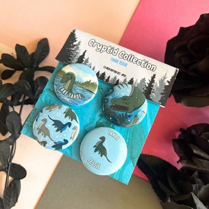 May include: A set of four 1.5 inch button pins featuring illustrations of the Lake Tahoe Tessie cryptid. The buttons are blue with white outlines and feature the text "Lake Tahoe Tessie" and "Cryptid Collection".
