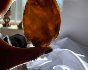 May include: A smooth, oval-shaped, brown agate stone with a translucent, cloudy appearance. The stone is held in a hand.