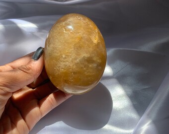 May include: A golden-yellow, egg-shaped crystal is held in a hand. The crystal has a smooth, polished surface with internal veining and a translucent quality. The background is a white, silky fabric.