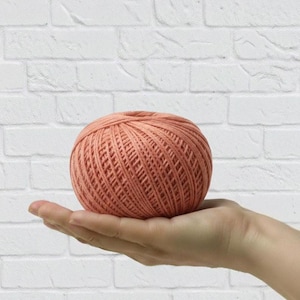 May include: A ball of peach-colored yarn, held in an open hand, against a white brick wall background. The yarn is tightly wound, showing a textured surface. Ideal for knitting or crochet projects.