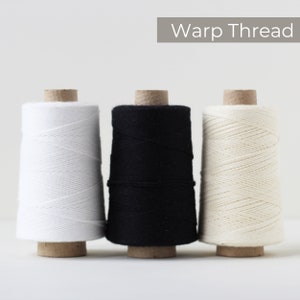 May include: Three spools of warp thread in white, black, and off-white. The spools are cylindrical, with the thread wound around a cardboard core. The text "Warp Thread" is displayed in a gray rectangle above the spools.