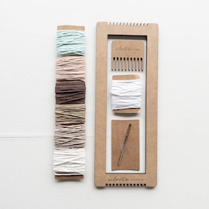 Op de afbeelding: Een knutselset met een houten frame, een kam, een naald en spoelen garen in verschillende kleuren. De garenkleuren omvatten tinten groen, roze, bruin en wit. De tekst "silento studio" staat op het frame.