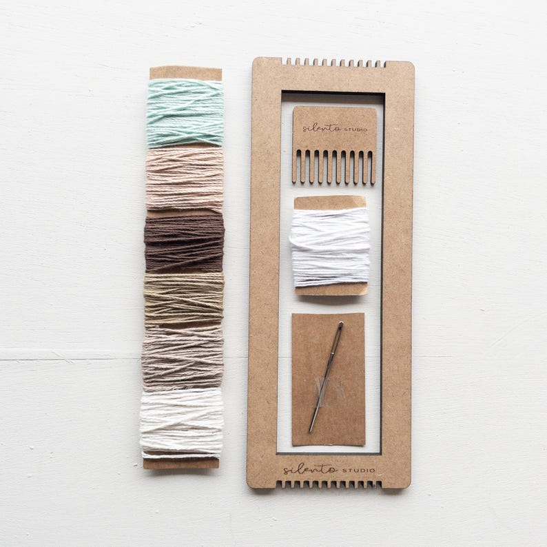 Op de afbeelding: Een knutselset met een houten frame, een kam, een naald en spoelen garen in verschillende kleuren. De garenkleuren omvatten tinten groen, roze, bruin en wit. De tekst "silento studio" staat op het frame.