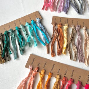 May include: An assortment of colorful silk ribbons in various shades, including pink, blue, green, orange, and red. Each ribbon is attached to a small, labeled cardboard tag. The ribbons are arranged on a white surface.