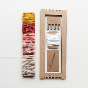 Op de afbeelding: Een knutselset met een houten weefgetouw, een kam, wit garen en een naald. Een stapel kleurrijk garen in tinten geel, rood en roze staat naast het weefgetouw. Op het weefgetouw staat de tekst "silento studio".