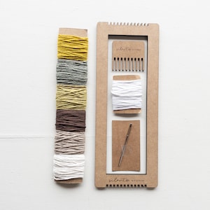 Op de afbeelding: Een weefset met een houten frame, een kam, een naald en spoelen garen in verschillende kleuren. De garenkleuren zijn geel, grijs, groen, bruin en wit. De tekst "silento studio" is op het frame gedrukt.