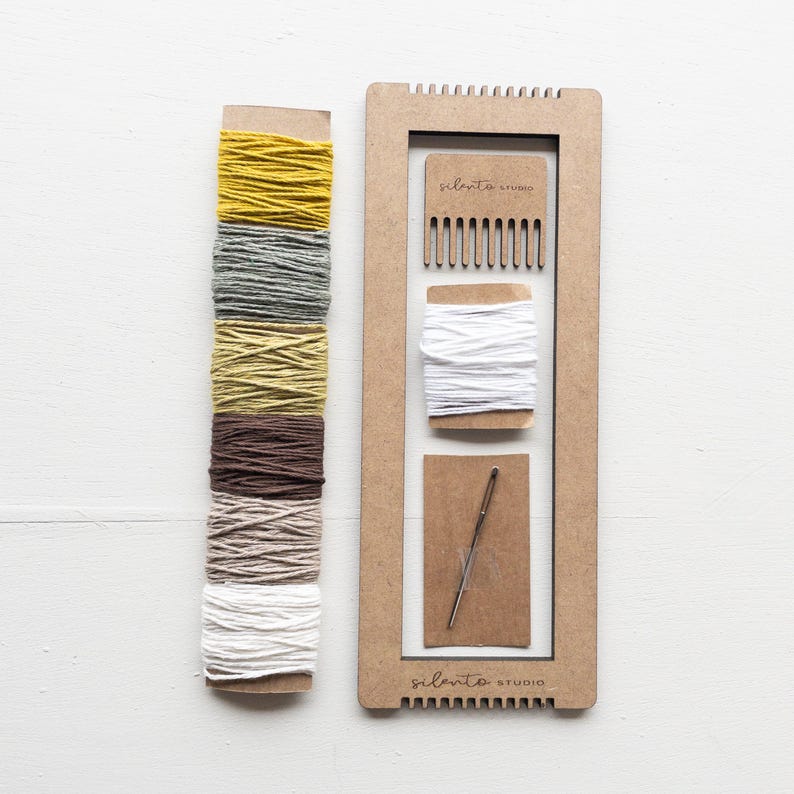 Op de afbeelding: Een weefset met een houten frame, een kam, een naald en spoelen garen in verschillende kleuren. De garenkleuren zijn geel, grijs, groen, bruin en wit. De tekst "silento studio" is op het frame gedrukt.