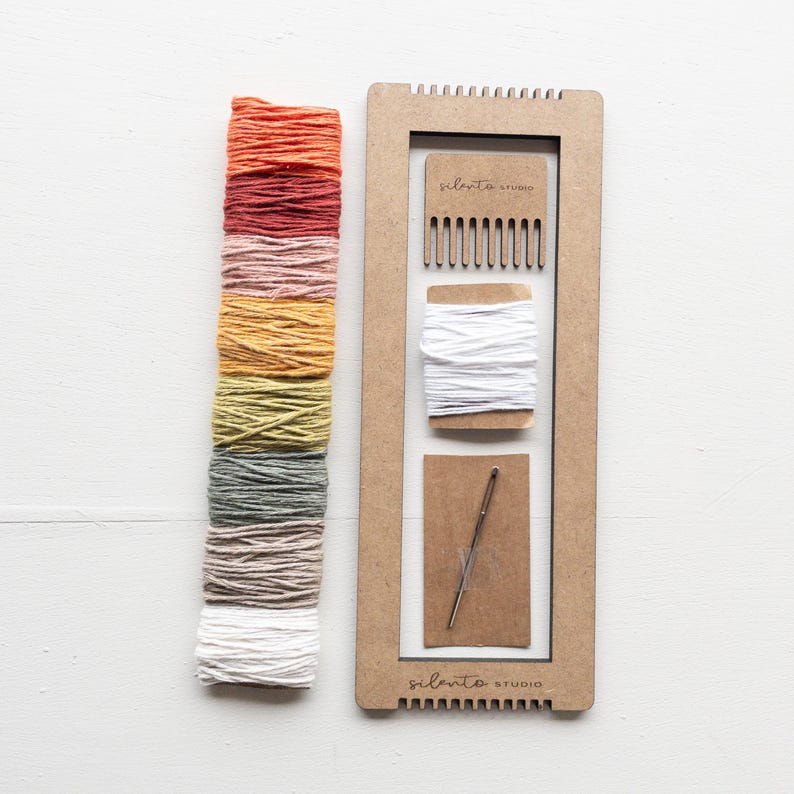 Op de afbeelding: Een weefset met een houten frame, kam, naald en witte draad. Een verloop van kleurrijk garen in tinten oranje, rood, roze, geel, groen en grijs wordt getoond. De tekst "silento studio" is zichtbaar op het frame.