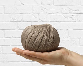Puede incluir: Una madeja de hilo color topo, sostenida en una mano abierta, sobre un fondo de pared de ladrillos blancos. El hilo parece estar bien enrollado, con hebras visibles. Adecuado para anuncios relacionados con la artesanía y el tejido.