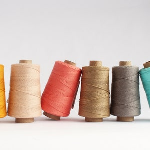 May include: Six spools of coloured thread in yellow, beige, coral, tan, grey, and teal. Each spool is wound around a cardboard core and leans slightly. The threads are tightly wound, creating a textured appearance.