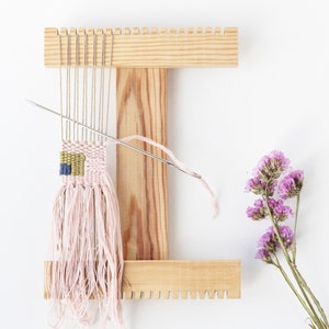 May include: A wooden loom with a pink and green woven tapestry hanging from it. A needle with pink thread is threaded through the loom. A bunch of dried purple flowers are on the right side of the image.