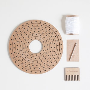 May include: A wooden circular loom with multiple concentric rings of holes, a white string, a wooden needle, a wooden comb with the text "silento studio", and a brown paper envelope.