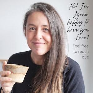 Op de afbeelding: Een vrouw met lang grijs haar glimlacht, terwijl ze een kleine, beige keramische kom vasthoudt. De afbeelding bevat de tekst "Hi! I'm Igone, happy to have you here! Feel free to reach out!"