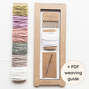Könnte beinhalten: Ein Webset mit einem Holzrahmen, einem Kamm, weißem Faden und einer Nadel. Das Set enthält eine Vielzahl von farbigen Garnen in Grün-, Lila-, Rosa- und Grautönen. Der Text "+ PDF weaving guide" ist ebenfalls sichtbar.