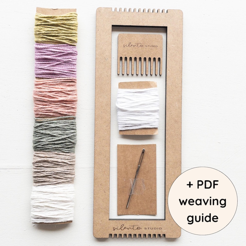 Op de afbeelding: Een weefset met een houten frame, kam, wit garen en een naald. De set bevat een verscheidenheid aan gekleurd garen in tinten groen, paars, roze en grijs. De tekst "+ PDF weaving guide" is ook zichtbaar.