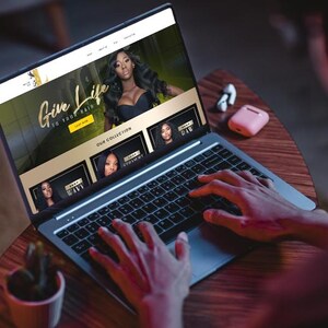 Puede incluir: Un port&aacute;til muestra un sitio web con el texto "Give Life to Your Hair" e im&aacute;genes de cabello largo y ondulado. El port&aacute;til est&aacute; sobre una mesa de madera, con una peque&ntilde;a planta y auriculares inal&aacute;mbricos. Manos escribiendo en el teclado.