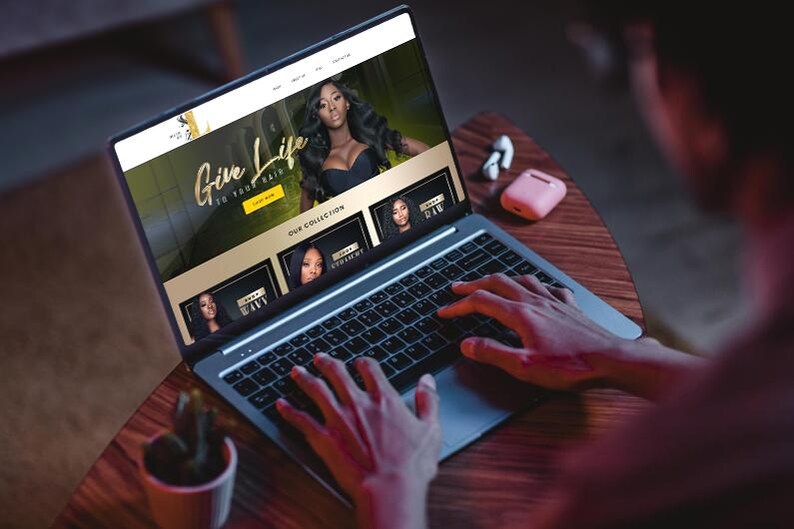 Puede incluir: Un port&aacute;til muestra un sitio web con el texto "Give Life to Your Hair" e im&aacute;genes de cabello largo y ondulado. El port&aacute;til est&aacute; sobre una mesa de madera, con una peque&ntilde;a planta y auriculares inal&aacute;mbricos. Manos escribiendo en el teclado.