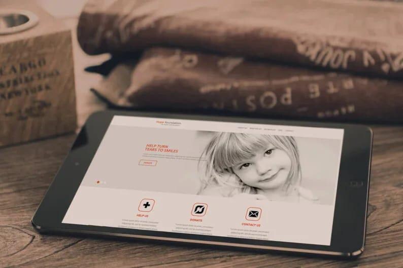 May include: A black tablet displays a website with a child's photo and the text "Help Turn Tears to Smiles." The tablet is on a wooden surface, with a wooden box and folded fabric in the background.
