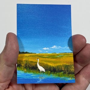 May include: Miniature painting of a coastal marsh scene. A white bird stands in the water, with yellow and green marsh grass in the background. The sky is blue with a few white clouds. The painting is on a small rectangular card.