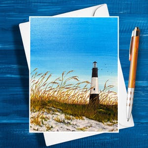 May include: A greeting card featuring a painting of a lighthouse on a sandy dune with tall grass. The lighthouse is black and white, set against a blue sky. A pen rests on the card.