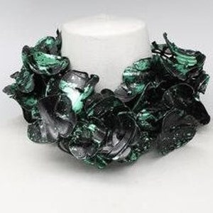 May include: A statement necklace featuring a cluster of dark green and black petal-shaped elements. The necklace is displayed on a white mannequin bust, showcasing its design and texture. The necklace appears to be made of a glossy material.
