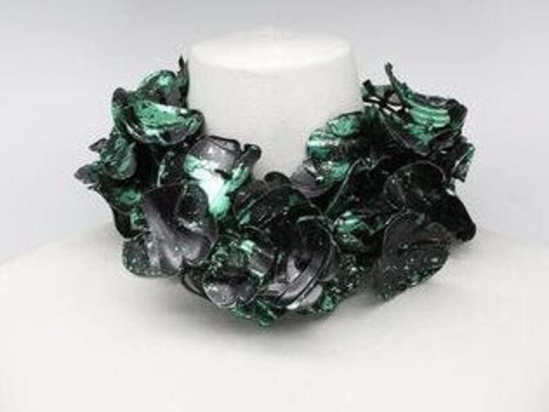 May include: A statement necklace featuring a cluster of dark green and black petal-shaped elements. The necklace is displayed on a white mannequin bust, showcasing its design and texture. The necklace appears to be made of a glossy material.